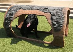 a black dog sitting in a wooden structure on the grass with his tongue hanging out