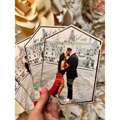 a hand holding up two wedding cards with the same photo on them and flowers in the background