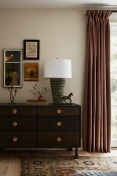 a room with a dresser, lamp and pictures on the wall next to it in front of a window