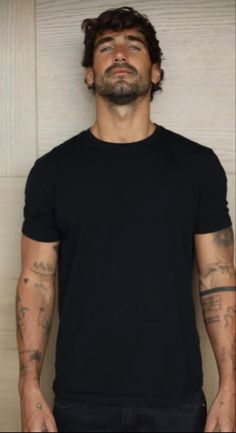 a man with tattoos standing in front of a white wall wearing a black t - shirt