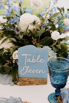 the table is set with blue and white flowers, greenery, and a sign that says table eleven