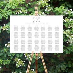 an easel with a seating chart on it in front of some bushes and white flowers