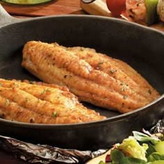 two fish fillets in a frying pan on a table with vegetables and bread