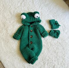 a green knitted frog outfit and matching booties laying on a white rug with the baby's hat