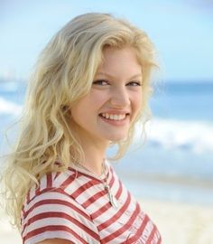 a beautiful blonde woman standing on top of a beach next to the ocean and smiling