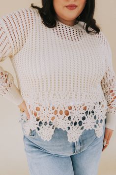 a woman wearing a white sweater with crochet on the shoulders and bottom, standing in front of a beige background