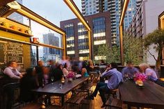 many people are sitting at tables outside in the evening with city lights and tall buildings behind them