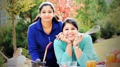 two women are posing for a photo in front of an outdoor table with food and utensils