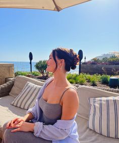 a woman sitting on top of a couch under an umbrella next to the ocean in front of her