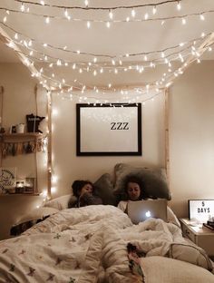 two people are laying in bed with their laptops under the covers and lights strung above them