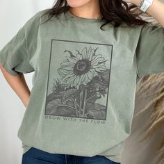 "This \"Grow With The Flow\" t-shirt is perfect for teachers, plant lovers, and anyone who wants to embrace the beauty of nature. The monochromatic sunflower design is pencil-drawn, giving it a vintage and retro feel. Made with Comfort Colors, this shirt is not only stylish, but also incredibly comfortable to wear. The botanical theme is perfect for garden and wildflower enthusiasts, and the floral tee makes a great addition to any wardrobe. Whether you're looking to inspire yourself or others, this shirt is a positive reminder to embrace the natural flow of life. Looking for a comfortable and durable t-shirt that you can customize to your liking? Look no further than Comfort Colors' garment-dyed tee! Here are some of the features that make this shirt a great addition to your wardrobe: -Ma Affordable Trendy Shirt With Plant Print, Green Cotton T-shirt With Plant Print, Green T-shirt With Plants Print, Relaxed Fit, Spring Crew Neck T-shirt For Outdoor, Green Cotton Shirt With Plant Print, Cotton Graphic Print T-shirt For Gardening, Cotton T-shirt With Graphic Print For Gardening, Green Cotton Tops With Plant Print, Green Cotton Top With Sunflower Print