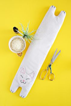 a cat pillow with scissors and other craft supplies on a bright yellow background, next to it is an air plant