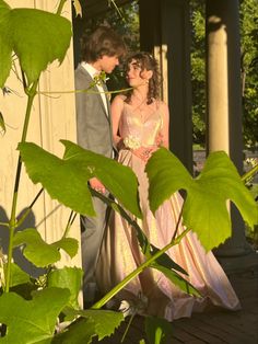 a man and woman standing next to each other in front of a house with large green leaves