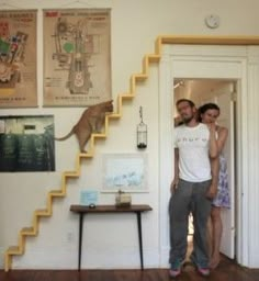 a man and woman standing in front of a stair case next to a table with a cat on it