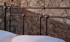 an iron bed frame with white pillows in front of a stone wall and lamp on the side