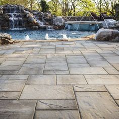 a stone patio next to a pool with waterfall in the back ground and water running down it