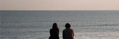 two people standing on the beach looking out at the ocean with a kite flying in the sky