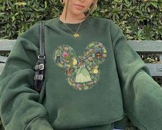 a woman sitting on a bench wearing a green sweatshirt with a mickey mouse head drawn on it