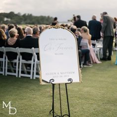 a sign that is sitting in the grass near some chairs and people standing around it