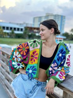 a woman sitting on a wooden bench next to the beach wearing a colorful crocheted jacket