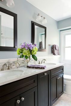 a bathroom with two sinks and mirrors on the wall next to it is purple flowers in a vase