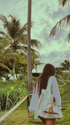 a woman walking down a path next to palm trees on a cloudy day with her back turned towards the camera