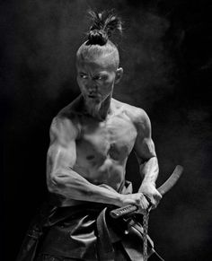 a shirtless man holding two swords in his hands and looking down at the camera