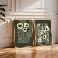 two framed art pieces sitting on top of a wooden floor next to a potted plant