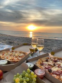 three pizzas and two glasses of wine are on the table near the ocean as the sun sets