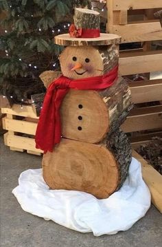 a snowman made out of logs sitting in front of a christmas tree with a red scarf around its neck