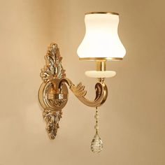 a gold wall light with a white shade on it's side and a crystal droplet hanging from the arm