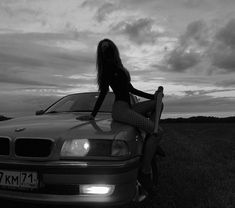 a woman sitting on the hood of a car in front of a field with clouds