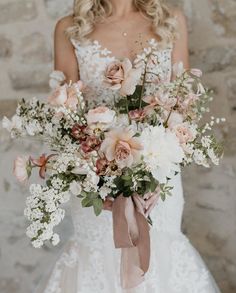 a bride holding a bouquet of white and pink flowers in front of a stone wall