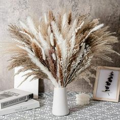 some dry grass in a white vase on a table next to two books and a framed photograph