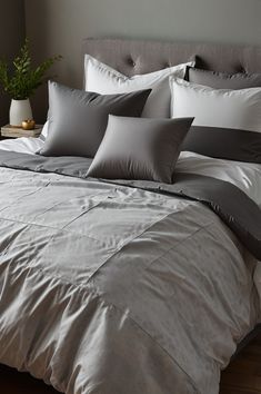 a bed with grey and white pillows on top of it next to a plant in a vase