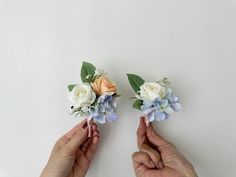 two hands holding flowers on top of a white surface with green leaves and pink and blue flowers