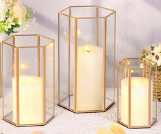 three gold lanterns with white candles in them on a table next to flowers and greenery