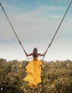 a woman in a yellow dress is swinging on a rope over some trees and bushes