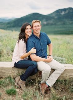 a man and woman sitting on a wooden bench in the grass with mountains in the background