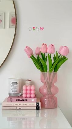 pink flowers are in a vase next to two books on a white table with a mirror