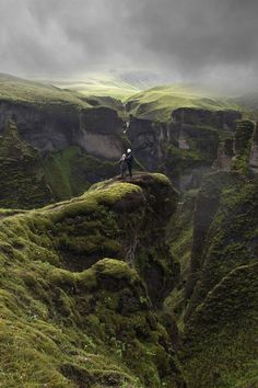 two people standing on the edge of a cliff with green mossy mountains in the background