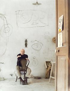 an older man sitting in a chair next to a white wall with drawings on it