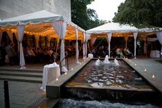 a group of people standing around a water fountain in front of white tents with lights on them