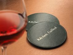 two black leather coasters with the words kitchen for food next to a glass of red wine