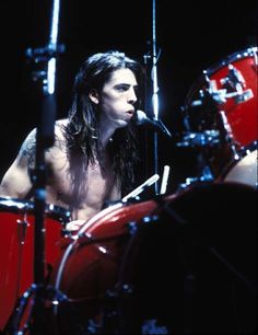 a man with long hair and no shirt on playing drums in front of a microphone