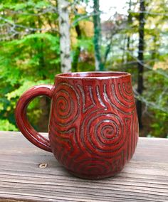 a red coffee mug sitting on top of a wooden table next to some green trees