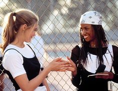 two young women standing next to each other near a chain link fence, talking and laughing