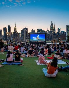 many people are sitting on the grass and watching movies in front of large city buildings