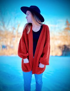 a woman standing in the snow wearing a red cardigan sweater and black floppy hat