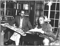 two men sitting at a table in front of a bookshelf and looking at papers